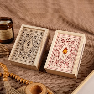 Pode incluir: Duas caixas de fósforos de madeira com desenhos florais e de tochas. Uma caixa apresenta um design gravado escuro, enquanto a outra tem um design impresso em vermelho e creme. As caixas retangulares são feitas de madeira clara.