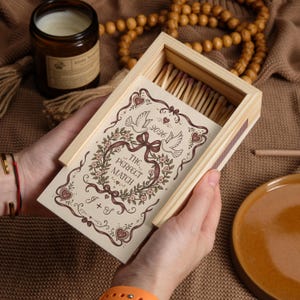 May include: A rectangular wooden matchbox containing wooden matches. The sliding cover features a brown ink illustration of two doves, a floral wreath, hearts, the text "THE PERFECT MATCH", "2026", and the initials "J + S" on a cream background.