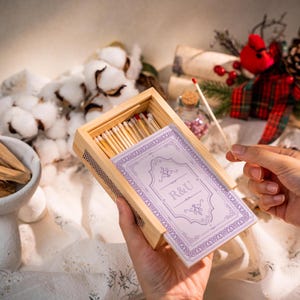 May include: A wooden matchbox with a decorative lavender label featuring the initials "R&U". The box is filled with wooden matches, some with red tips. The matchbox is held in a person's hands.