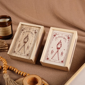 May include: Two rectangular wooden matchboxes featuring a graphic of two crossed lit matches, the text "THE PERFECT MATCH", "EST. 2026", and the letters "D" and "R". One box has a natural wood grain finish, while the other has a white lid with red ink.