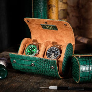 May include: A green, crocodile-textured watch case with two compartments, each holding a wristwatch. The interior is tan suede. The case is open, revealing the watches. The initials "MB" are embossed on the inside of the lid. A small, matching case is visible.