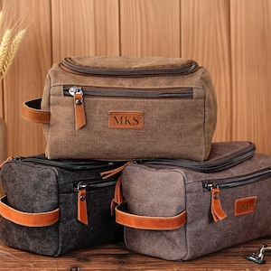 May include: Three canvas toiletry bags in brown, black, and gray, each with a zippered top, leather accents, and a handle. The brown bag has "MKS" on a leather patch.