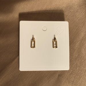 Puede incluir: Un par de pendientes de color dorado con un diseño rectangular simple. Los pendientes se muestran en una tarjeta blanca con un pequeño lazo.