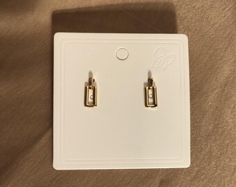 May include: A pair of gold-toned earrings with a simple rectangular design. The earrings are displayed on a white card with a small bow.