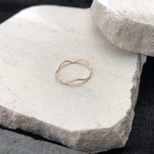 Puede incluir: Un delicado anillo de oro con un diseño ondulado y orgánico. El anillo se encuentra sobre una superficie de piedra blanca, mostrando sus intrincados detalles y sutil textura. El diseño del anillo recuerda a una ola fluida.