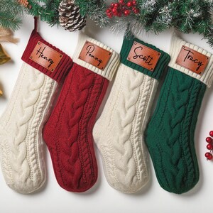 May include: Four cable-knit Christmas stockings in cream, red, and green, each with a personalized leather name tag. Festive decor includes pine boughs, berries, and gold bells, creating a cozy holiday scene.