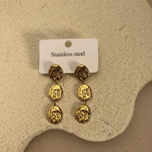 May include: Gold-toned stainless steel earrings with three round, textured, dangling charms. The earrings are displayed on a white card with the text "Stainless steel".