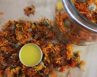May include: A small jar filled with dried orange and yellow flower petals. A small round container with a yellow substance is in front of the jar.
