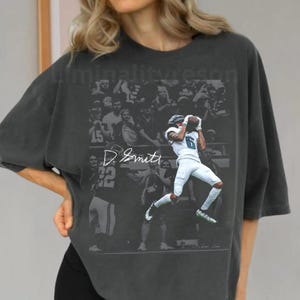 May include: Dark gray oversized t-shirt featuring a graphic of a football player in action, catching a ball. The shirt has a vintage, faded look with the player's name and number visible. The shirt is worn with black shorts.