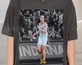 May include: Dark gray t-shirt featuring a black and white image of a basketball player in action. The player is wearing a white uniform with blue and gold accents. The word "INDIANA" is visible on the court.