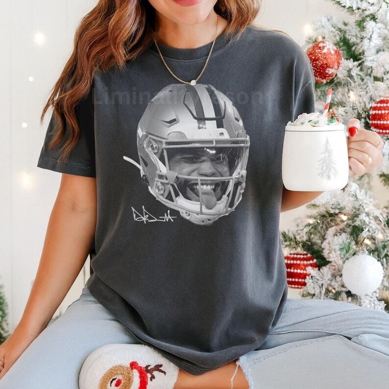 Puede incluir: Camiseta gris oscuro con una imagen en blanco y negro de un casco y la cara de un jugador de f&uacute;tbol americano. La camiseta tambi&eacute;n presenta la firma del jugador. La persona lleva una taza con un dise&ntilde;o de &aacute;rbol de Navidad.