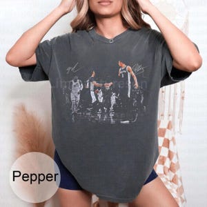 May include: Dark gray t-shirt featuring a basketball game scene with player signatures. The shirt has a crew neck and short sleeves. The word "Pepper" is printed on a white oval in the bottom left corner.