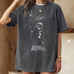 May include: Dark gray t-shirt featuring a black and white image of a football player raising his arm. The player is wearing a helmet and jersey with the number 1. The shirt has the text "Liminality" and a signature.