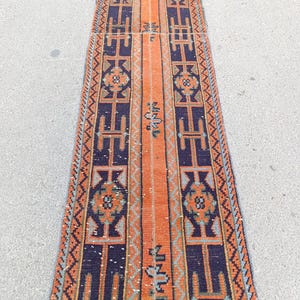 May include: A long, narrow runner rug with a central orange stripe flanked by dark blue panels. The panels feature geometric patterns in orange, blue, and white. The rug has a vintage, handcrafted appearance, suitable for hallways or entryways.
