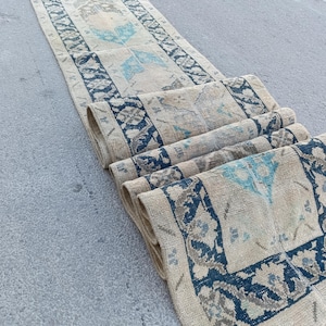 May include: A long, narrow runner rug featuring a cream-coloured base with a repeating geometric pattern in dark navy blue and accents of bright turquoise. The rug has a wide, decorative border and is partially folded to show its woven texture.