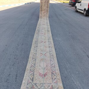 May include: An exceptionally long, narrow runner rug featuring a faded, distressed aesthetic with a neutral beige base. The rug is decorated with intricate, repeating geometric and floral patterns in muted shades of red, blue, and soft green.