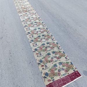 May include: A long, narrow runner rug featuring a cream-coloured base with a repeating floral pattern in shades of pink, blue, and yellow. The rug is finished with solid deep red bands at both ends and has a short, light-coloured fringe at the edges.