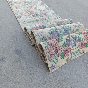 Pode incluir: Um tapete passadeira longo e estreito com uma base de cor creme e um padrão floral repetitivo em tons de rosa, azul e verde. O tapete está parcialmente enrolado numa extremidade, revelando a textura tecida e o intrincado design botânico multicolorido.