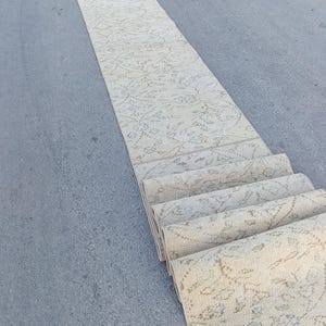 May include: A long, narrow runner rug featuring a light beige base color with a faded, intricate floral pattern in muted gray and tan tones. The rug is partially rolled at one end, displaying a repeating botanical design across its surface.