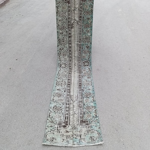 Pode incluir: Um tapete longo e retangular com um intrincado padrão floral e geométrico. O tapete apresenta uma paleta de cores desbotadas de verde-azulado, castanho e creme. O design inclui uma coluna central e bordas ornamentadas, criando uma estética vintage. O tapete é segurado por uma pessoa.