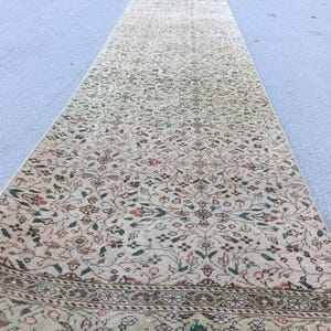 May include: A long, narrow runner rug featuring a beige base with an intricate, repeating floral pattern in muted shades of green, red, and brown. The rug has a detailed decorative border along the bottom edge with similar floral motifs.