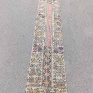 May include: A long, rectangular rug with a central pink stripe and intricate floral patterns in shades of pink, yellow, gray, and green. The rug has a faded, vintage appearance, suggesting it is handmade.