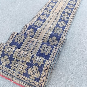 May include: A long, narrow runner rug featuring a deep blue and beige color palette. The design consists of repeating geometric star-like motifs arranged in a central column, bordered by intricate linear patterns and a red fringe at the end.