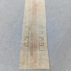 May include: A rectangular, vintage-style rug with a faded, distressed appearance. The rug features a central stripe of light brown and beige tones, flanked by patterned borders in similar hues. The rug's design suggests a traditional, handcrafted aesthetic.