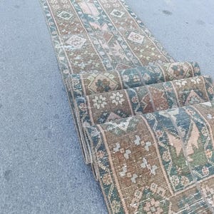 May include: A long, narrow runner rug featuring a repeating geometric pattern in muted shades of brown, sage green, and soft pink. The textile displays a series of interconnected diamond and floral motifs arranged in vertical bands across the surface.