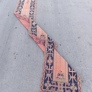 Peut inclure: Un long tapis de couloir étroit présentant un champ de couleur pêche délavée avec des bordures géométriques bleu marine foncé et des motifs centraux. Le tapis affiche un motif répétitif de formes angulaires et de petits éléments floraux.