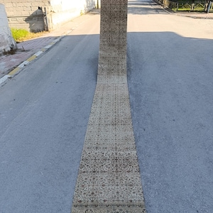 May include: A long, rectangular, vintage rug with an intricate floral pattern in shades of beige, brown, and burgundy. The rug is displayed outdoors on a gray asphalt surface, showcasing its length and design.