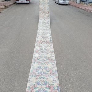 Puede incluir: Una alfombra larga y rectangular con un estampado floral en tonos rosa, azul y verde sobre un fondo crema. La alfombra se exhibe al aire libre sobre una superficie pavimentada, mostrando su longitud y diseño.