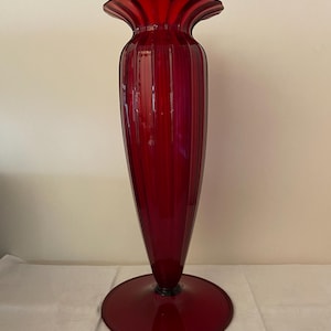 Puede incluir: Un jarrón alto de cristal rojo rubí con la parte superior acampanada y una base circular ancha. El jarrón tiene detalles verticales acanalados y un cuerpo cónico y esbelto. La base mide unos 15 cm de diámetro.