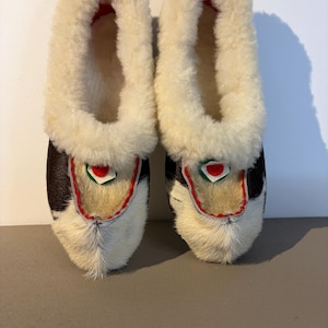 Op de afbeelding: Een paar met bont gevoerde pantoffels. De pantoffels hebben een witte bontvoering en een zwart-wit patroon aan de buitenkant. Het ontwerp bevat een rode en groene accentkleur bij de teen. Deze pantoffels zijn voor binnenshuis.