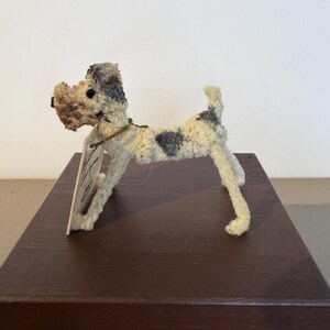 May include: A handmade terrier dog figurine with a cream-colored body and gray spots. The dog has a textured, fuzzy appearance and a small tag attached to its neck. It is posed on a dark brown surface.