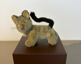 Puede incluir: Un juguete de gato de peluche vintage con un cuerpo marrón y gris, cola negra y ojos verdes. El gato está posado sobre un bloque de madera marrón. El gato tiene un pequeño lazo amarillo alrededor del cuello.