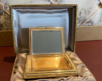 May include: A gold-colored compact mirror in a light gray velvet box with a gold trim. The mirror is open and the inside is lined with a light gray fabric.
