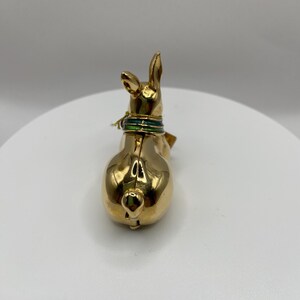 以下が含まれることがあります： 金色のウサギの置物。緑と金の首輪が付いています。ウサギは背中を観客に向けており、丸みを帯びた形と小さな尻尾を見せています。置物は白い表面に置かれています。