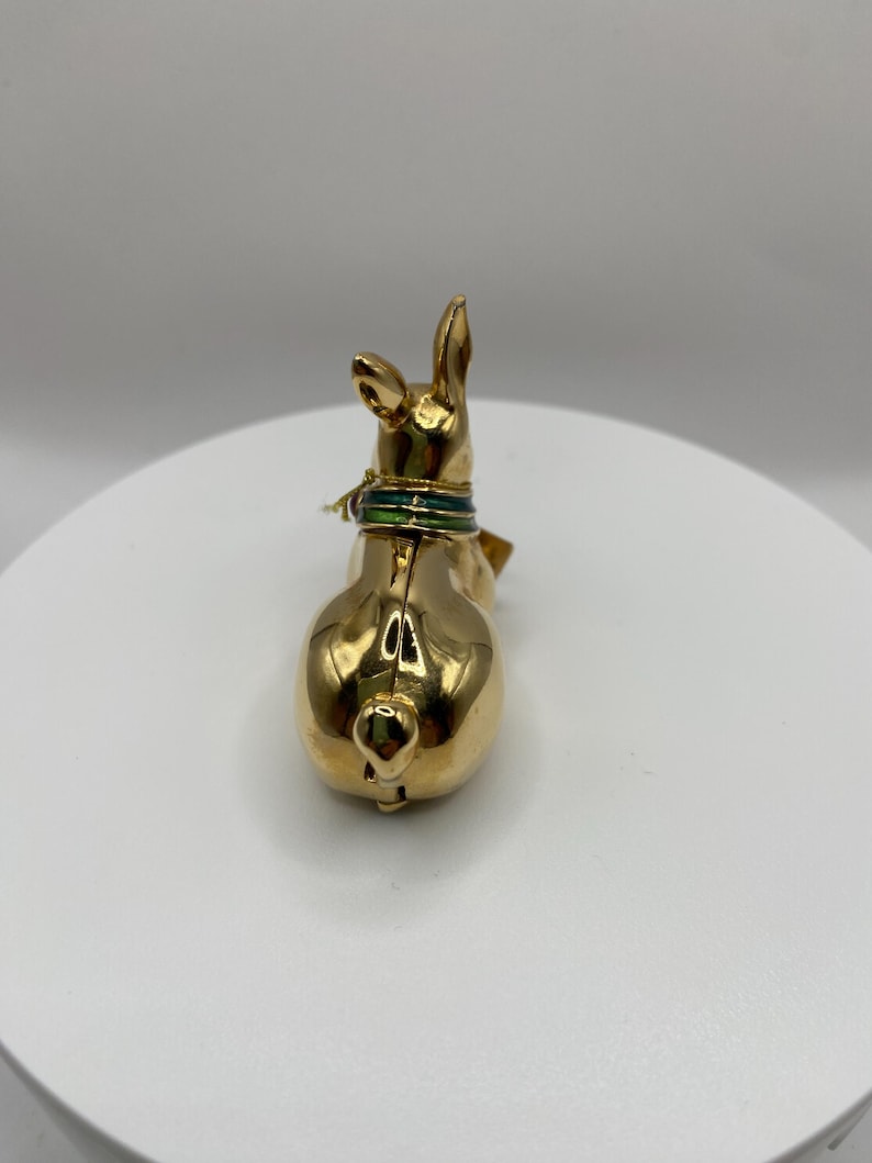 以下が含まれることがあります： 金色のウサギの置物。緑と金の首輪が付いています。ウサギは背中を観客に向けており、丸みを帯びた形と小さな尻尾を見せています。置物は白い表面に置かれています。