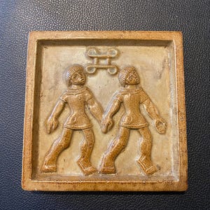 Peut inclure: Carreau de céramique carré, de couleur beige, représentant en relief deux personnages se tenant la main, avec un symbole au-dessus. Les personnages portent des tuniques. Le carreau a un bord en relief.