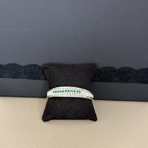 以下が含まれることがあります： 小さな丸い緑色の石と長方形の白い石が並んだシルバーブレスレット。 ブレスレットは、ニュートラルな背景に対して黒いクッションの上に表示されています。 ジュエリーはバングルスタイルです。