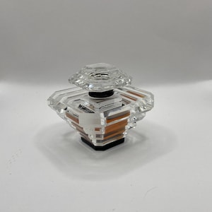 Op de afbeelding: Een heldere, facetgeslepen parfumfles met een geometrisch ontwerp. De fles heeft een doorzichtige, diamantvormige stopper en een zwarte basis. De vloeistof binnenin is amberkleurig. De fles staat op een witte ondergrond.