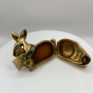 以下が含まれることがあります： 金色のウサギ型の香水コンパクトで、蓋が開いています。ウサギはピンク色の宝石の目と緑と紫の襟をしています。コンパクトには茶色の固形香水が入っています。小さなタグがウサギに付いています。