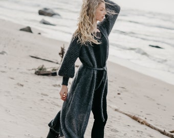 Könnte beinhalten: Eine Frau trägt einen langen, grauen Cardigan mit einem schwarzen Oberteil und einer schwarzen Hose. Sie steht an einem Sandstrand mit dem Meer im Hintergrund. Sie trägt schwarze Stiefel mit Schnürsenkeln.