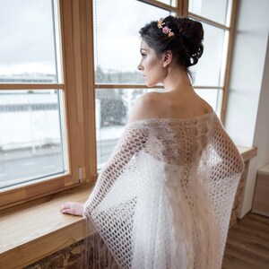 May include: A woman in a white dress with a delicate lace shawl, standing by a window. She is wearing a floral hairpiece and looking out the window.