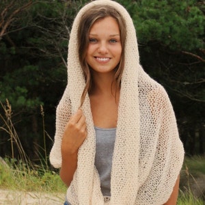 May include: A cream-coloured, open-weave hooded scarf is draped around the shoulders. The scarf is made of a soft, lightweight material. The person is wearing a grey vest top and blue denim shorts. The background is a blurred view of green trees.
