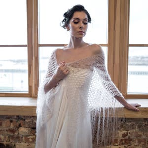 May include: A woman wearing a white crocheted shawl with a delicate pattern and long fringe. The shawl is draped over a white dress and the woman is standing in front of a window.