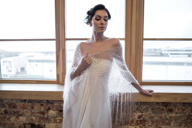 May include: A woman wearing a white crocheted shawl with a delicate pattern and long fringe. The shawl is draped over a white dress and the woman is standing in front of a window.