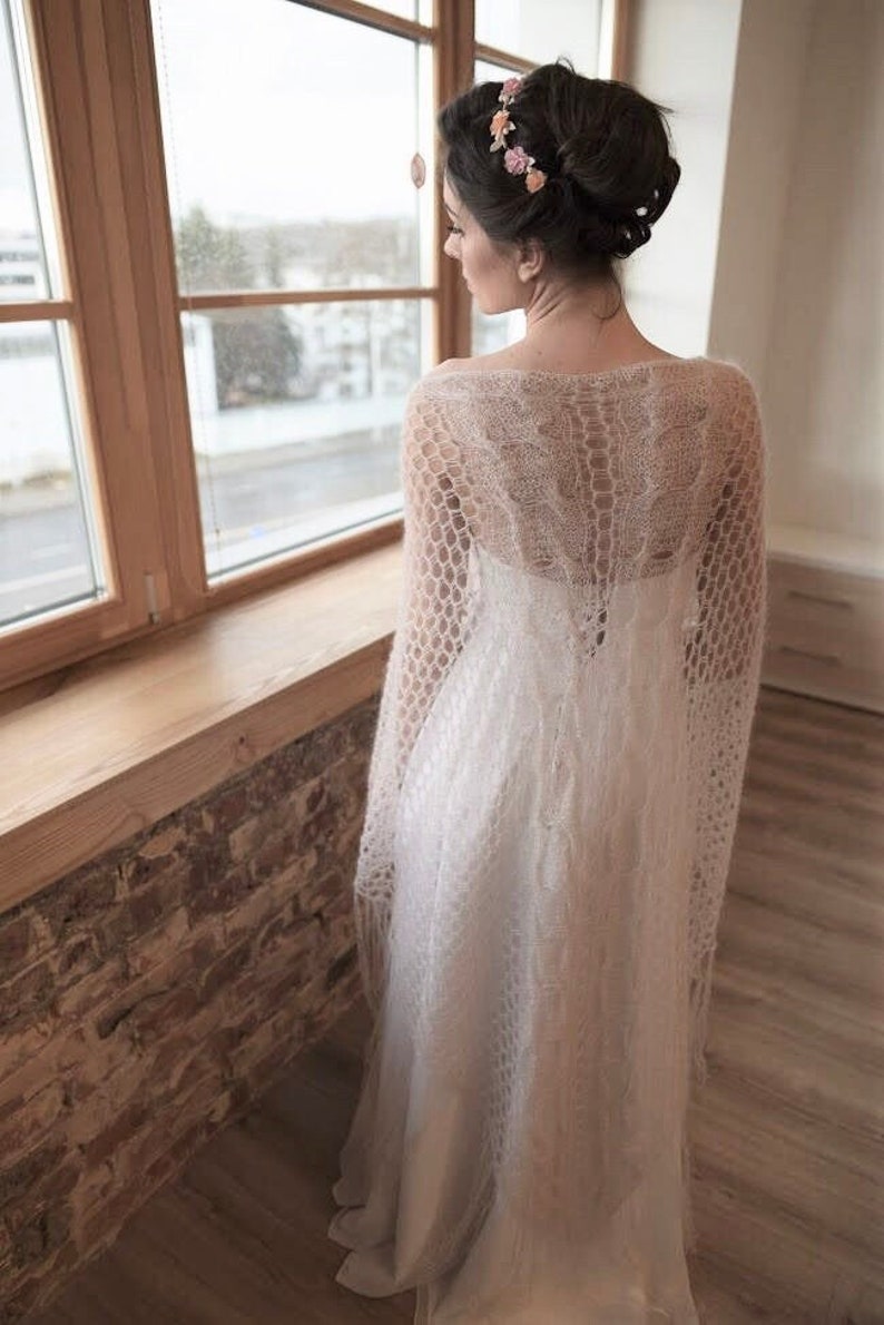 May include: A white knitted shawl with a delicate openwork pattern. The shawl is draped over a person wearing a white dress and a floral crown.