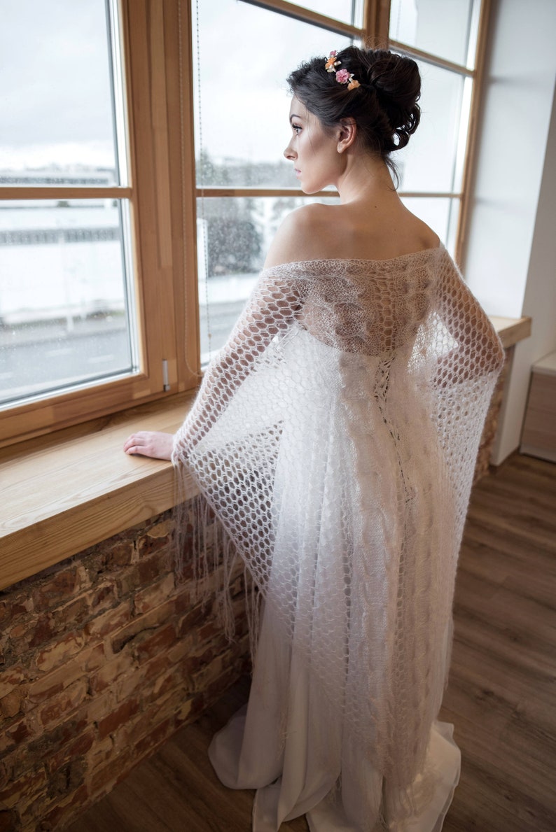May include: A woman in a white dress with a delicate lace shawl, standing by a window. She is wearing a floral hairpiece and looking out the window.